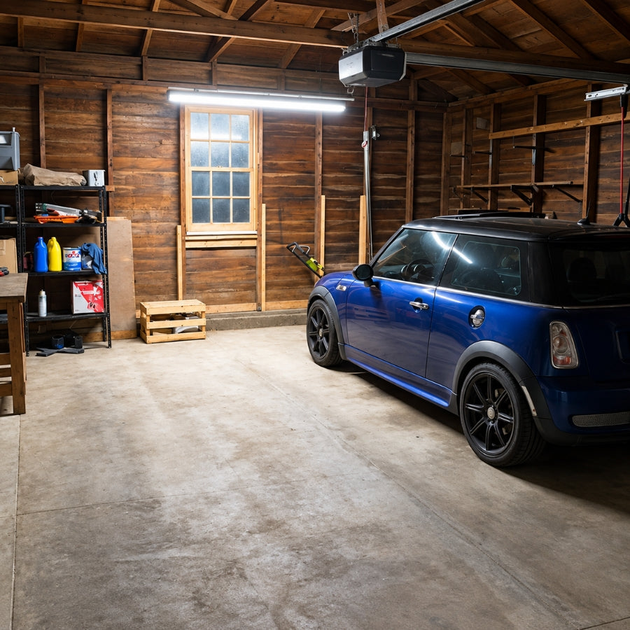A blue Mini Cooper is parked inside a rustic wooden garage with concrete floors, shelving, and a window.